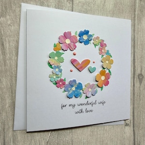 May include: A white greeting card with a floral wreath design. The wreath is composed of colorful, layered paper flowers in shades of pink, blue, yellow, and green. Two hearts and the text "for my wonderful wife with love" are in the center.