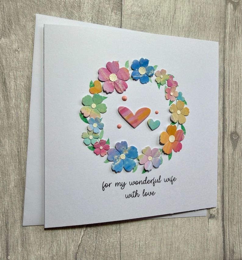 May include: A white greeting card with a floral wreath design. The wreath is composed of colorful, layered paper flowers in shades of pink, blue, yellow, and green. Two hearts and the text "for my wonderful wife with love" are in the center.