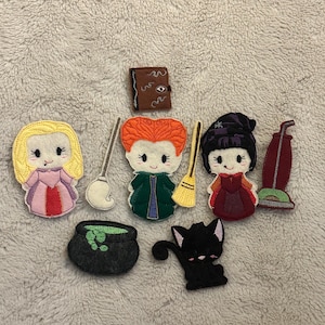 May include: A collection of felt appliques featuring characters and items from a Halloween-themed movie. Includes figures of witches, a black cat, a cauldron, a spell book, a vacuum cleaner, and brooms. The appliques are in various colors, including pink, green, and black.