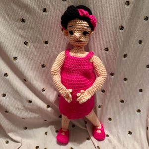 May include: A crocheted doll wearing a pink dress and pink shoes. The doll has black hair and a pink headband.