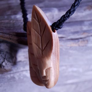 Può includere: Un ciondolo in legno scolpito con un design a foglia e volto, sospeso a un cordino nero. Il ciondolo è marrone chiaro con venature del legno visibili e intaglio dettagliato. Il cordino è in materiale nero intrecciato.