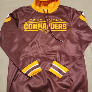 May include: A maroon hoodie with a yellow and white striped trim. The hoodie has a yellow hood and a white drawstring. The front of the hoodie has the words "WASHINGTON COMMANDERS" in yellow letters with a white "W" in the center.