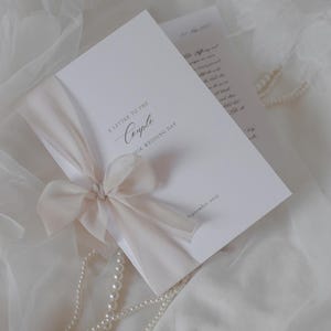 Könnte beinhalten: Eine weiße Hochzeitskarte mit dem Text "A Letter to the Couple" und "Their Wedding Day" in eleganter Schrift. Die Karte ist mit einem cremefarbenen Band und einer Schleife gebunden. Eine Perlenkette und eine zweite Karte mit handschriftlichem Text sind ebenfalls zu sehen.