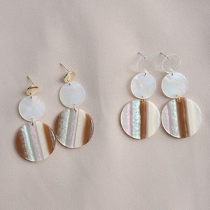 May include: Two pairs of dangle earrings with a layered circular design. Each earring features a small gold or clear top, a white iridescent middle circle, and a larger bottom circle with brown and white stripes. The earrings are on a neutral background.