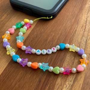 May include: A colorful beaded phone charm with the name "ALEX" spelled out in white letters. The charm is made with white, purple, orange, yellow, green, and blue beads in the shape of hearts and stars.