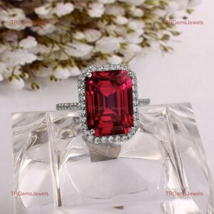 Puede incluir: Un anillo de tono plateado con una gran piedra preciosa roja rectangular de corte esmeralda en el centro, rodeada por un halo de pequeñas piedras claras de corte redondo. La banda es delgada y también tiene piedras claras engastadas.