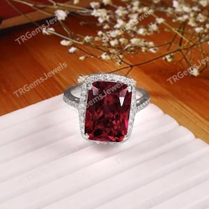 May include: A silver-toned ring featuring a large, rectangular, deep red gemstone set in the centre. The gemstone is surrounded by a halo of small, clear, sparkling stones, with additional small stones embedded along the band of the ring.