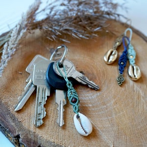 Könnte beinhalten: Ein Schlüsselsatz mit einem grünen und weißen Makramee-Schlüsselanhänger mit einer kleinen weißen Muschel. Zwei weitere Makramee-Schlüsselanhänger mit Muscheln sind ebenfalls abgebildet, einer in Blau und einer in Grün.