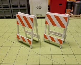 May include: Two white plastic construction barricades with orange and white striped panels. The barricades are folded and standing on a green grid surface.