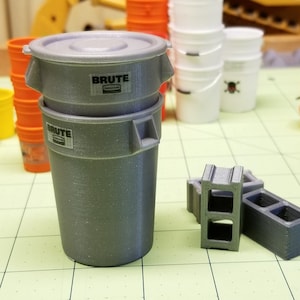 May include: Two gray plastic trash cans with the word "BRUTE" printed on the side. The cans are stacked on top of each other. There are also two gray concrete blocks in the image.
