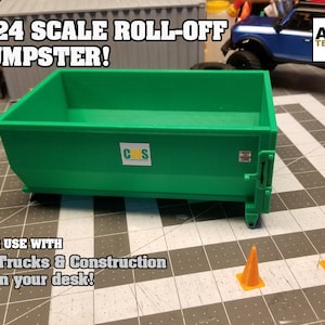May include: A green 1/24 scale roll-off dumpster model with a "CWS" logo. The dumpster is on a checkered surface with two orange traffic cones.
