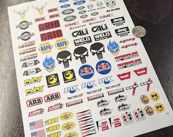 Peut inclure: Une feuille d'autocollants colorés représentant divers logos et marques, notamment Ford, Jeep, Ram et Red Bull. Les autocollants sont conçus pour les crawlers RC et sont imperméables. Le texte "RC Crawler Decals!" est en haut de la feuille. Le texte "POLYESTER MATERIAL! WATERPROOF! TRIM TO FIT/PEEL/APPLY!" est en bas de la feuille.