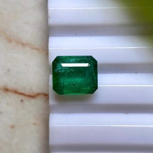 Peut inclure: Une pierre précieuse émeraude rectangulaire avec une teinte vert vif et une taille à facettes. La pierre présente des inclusions naturelles internes et une surface polie, reflétant la lumière sur ses facettes supérieures sur une base blanche.