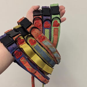 Könnte beinhalten: Eine Auswahl an bunten Hundehalsbändern mit schwarzen Kunststoffschnallen. Die Halsbänder weisen verschiedene Webmuster in Rot-, Blau-, Gelb- und Grüntönen auf. Jedes Halsband hat einen roten herzförmigen Aufnäher mit einem Logo.