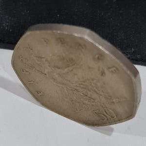 Puede incluir: Una moneda de color plateado con una forma inusual, no circular. La moneda presenta un dise&ntilde;o en relieve y el n&uacute;mero "50" en su superficie. La moneda est&aacute; ligeramente inclinada, revelando su borde y los detalles del dise&ntilde;o.