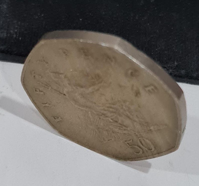 Puede incluir: Una moneda de color plateado con una forma inusual, no circular. La moneda presenta un dise&ntilde;o en relieve y el n&uacute;mero "50" en su superficie. La moneda est&aacute; ligeramente inclinada, revelando su borde y los detalles del dise&ntilde;o.