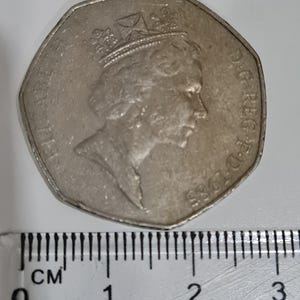 Puede incluir: Una moneda plateada de cincuenta peniques con el perfil de la reina Isabel II, fechada en 1985. La moneda est&aacute; junto a una regla con medidas en cent&iacute;metros. El di&aacute;metro es de aproximadamente 2,7 cm.