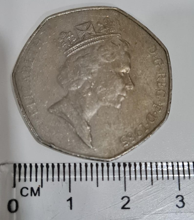 Puede incluir: Una moneda plateada de cincuenta peniques con el perfil de la reina Isabel II, fechada en 1985. La moneda est&aacute; junto a una regla con medidas en cent&iacute;metros. El di&aacute;metro es de aproximadamente 2,7 cm.