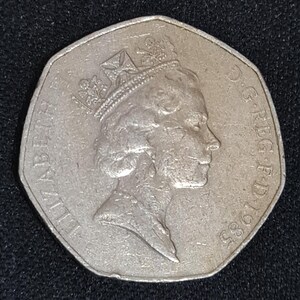 Puede incluir: Una moneda plateada de cincuenta peniques de 1985, con un perfil de la reina Isabel II con corona. La moneda tiene una forma heptagonal distintiva y la inscripci&oacute;n "ELIZABETH II D.G. REG. F.D. 1985" alrededor del borde.