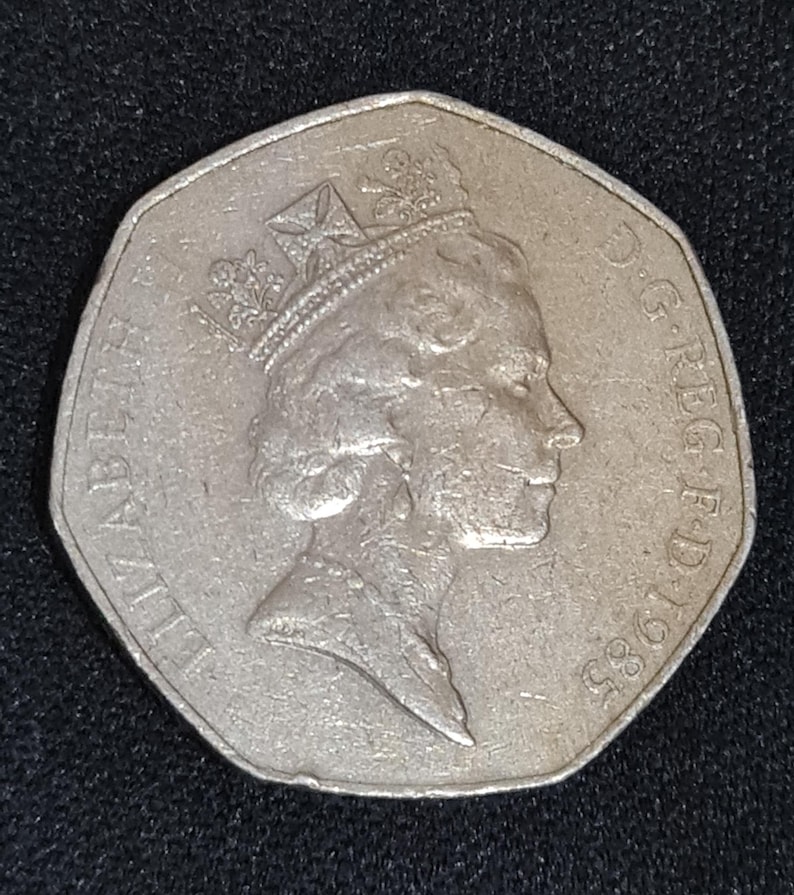 Puede incluir: Una moneda plateada de cincuenta peniques de 1985, con un perfil de la reina Isabel II con corona. La moneda tiene una forma heptagonal distintiva y la inscripci&oacute;n "ELIZABETH II D.G. REG. F.D. 1985" alrededor del borde.