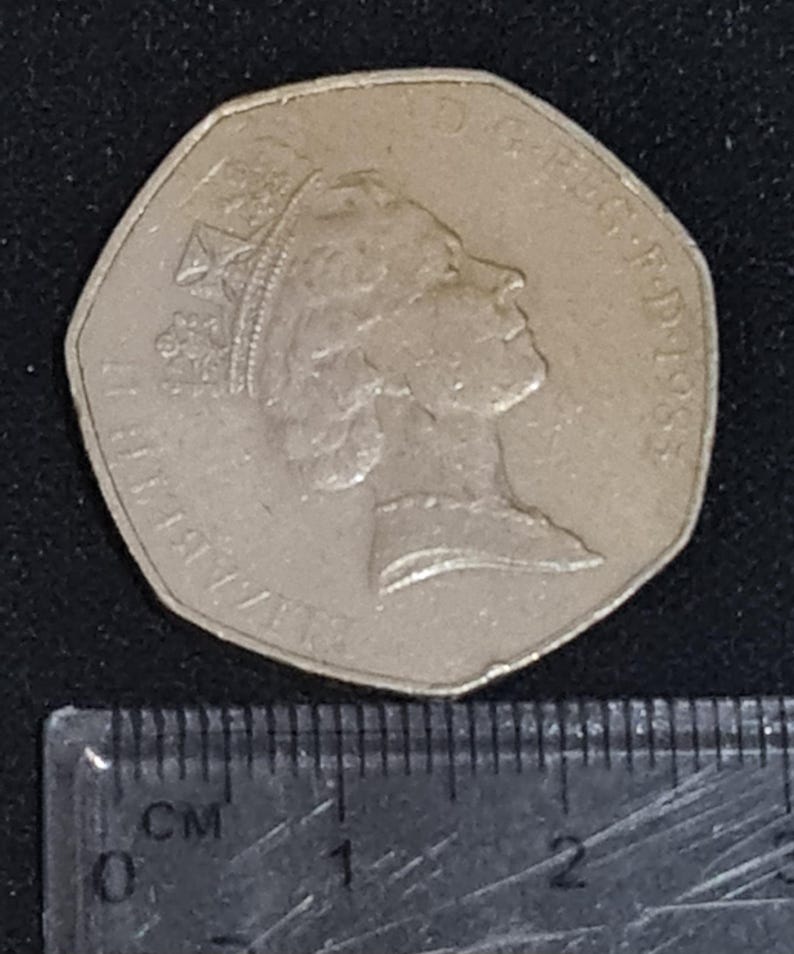 Puede incluir: Una moneda de cincuenta peniques de plata con el perfil de la reina Isabel II, con el a&ntilde;o 1983 visible. La moneda est&aacute; colocada junto a una regla con marcas de cent&iacute;metros.