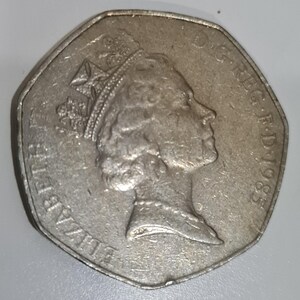 Puede incluir: Una moneda de cincuenta peniques de plata, de forma octogonal, que representa el perfil de la reina Isabel II, con una corona. La moneda lleva la inscripci&oacute;n "ELIZABETH II D.G.REG.F.D.1985" en el borde. La moneda est&aacute; ligeramente desgastada.