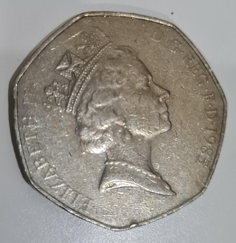 Puede incluir: Una moneda de cincuenta peniques de plata, de forma octogonal, que representa el perfil de la reina Isabel II, con una corona. La moneda lleva la inscripci&oacute;n "ELIZABETH II D.G.REG.F.D.1985" en el borde. La moneda est&aacute; ligeramente desgastada.