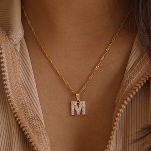 May include: A gold chain necklace with a pendant in the shape of the letter M. The pendant is encrusted with small, clear rhinestones.