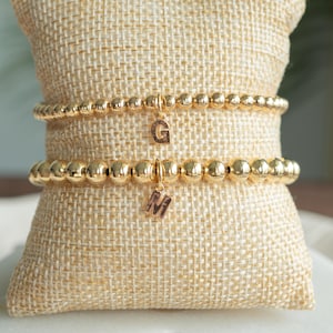May include: Two gold-toned beaded stretch bracelets featuring polished spherical beads. Each bracelet includes a single gold-toned letter charm hanging from the center, with one charm displaying the letter G and the other displaying the letter M.