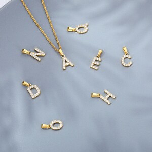 May include: A gold chain necklace with a variety of letter pendants, each letter is encrusted with clear rhinestones. The letters include A, C, D, E, H, N, and O.