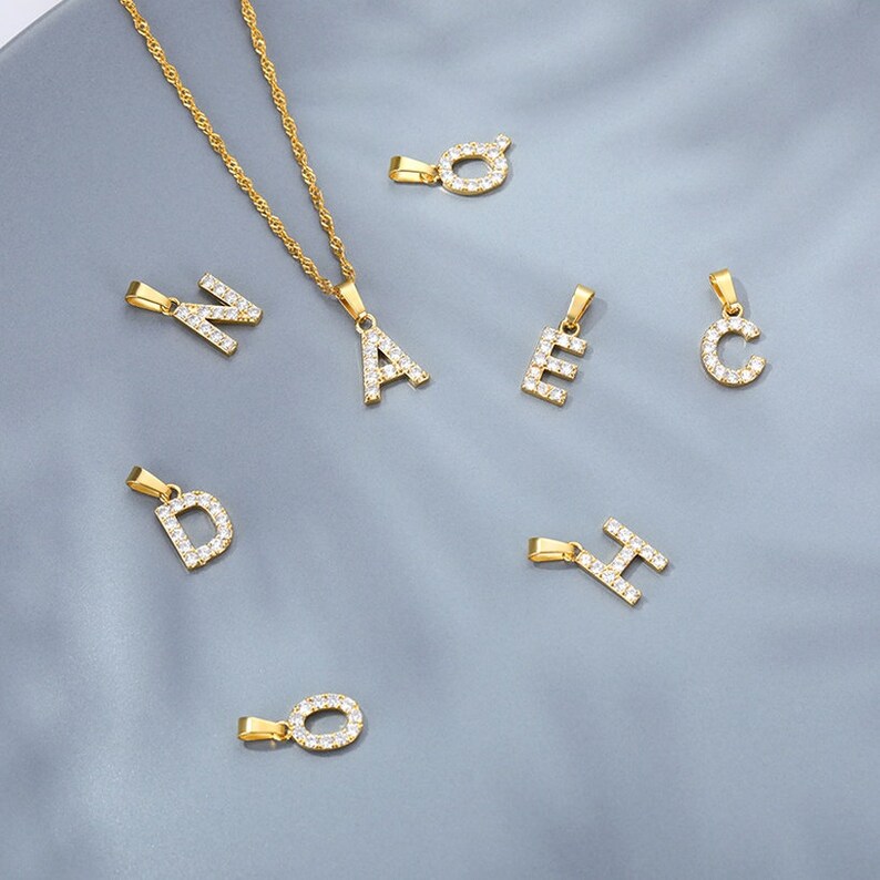 May include: A gold chain necklace with a variety of letter pendants, each letter is encrusted with clear rhinestones. The letters include A, C, D, E, H, N, and O.