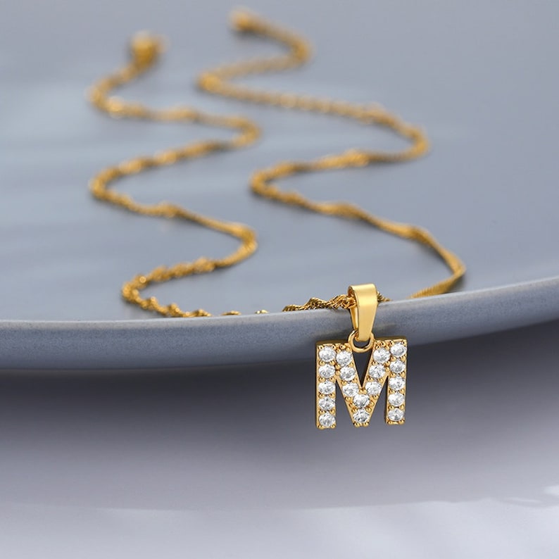 May include: Gold chain necklace with a pendant in the shape of the letter M. The pendant is encrusted with clear cubic zirconia stones.