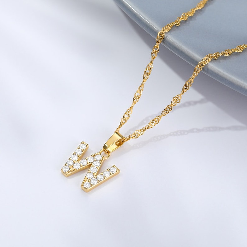 May include: Gold chain necklace with a pendant in the shape of the letter W, encrusted with clear cubic zirconia stones.