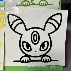 May include: A black and white vinyl decal featuring a stylized cartoon creature with long ears, pointed markings, and a circular forehead emblem. The character is depicted peeking over a horizontal line with its two front paws visible.