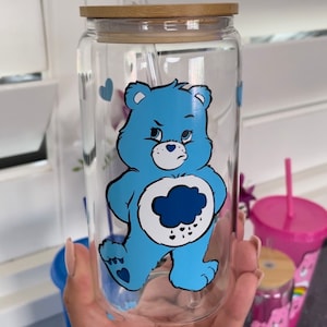 May include: A clear glass tumbler with a bamboo lid and a clear glass straw. The glass features a graphic of a light blue bear with a dark blue rain cloud symbol on its white belly, surrounded by small blue hearts printed on the exterior surface.