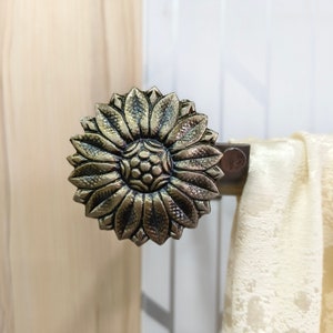 May include: A gold metal curtain tieback with a sunflower design. The tieback is attached to a curtain rod.