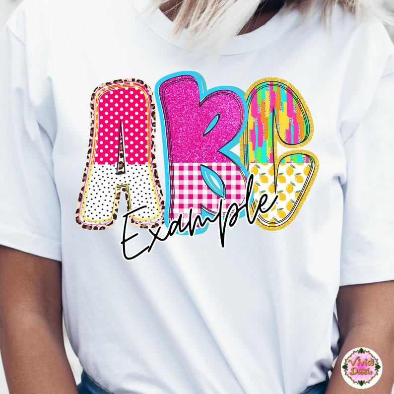 May include: White t-shirt featuring the letters "ABC" in a colorful, patterned design. The letter "A" has a pink polka dot and leopard print pattern. The letter "B" is pink glitter with a blue border. The word "Example" is written below.