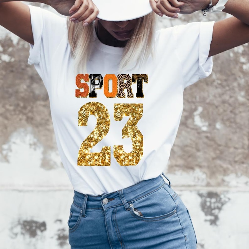 May include: White t-shirt with a gold glitter "SPORT 23" design. The letters are orange, black, and white.