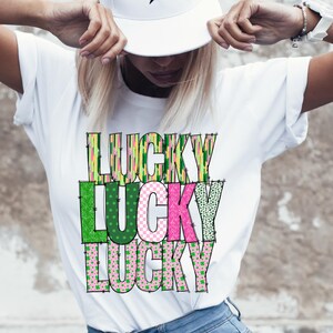 Pu&ograve; includere: Maglietta bianca con la parola "LUCKY" ripetuta tre volte in un design impilato. Ogni lettera &egrave; composta da diversi tessuti a motivi verdi, rosa e gialli.