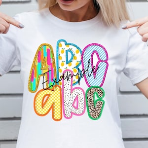 May include: White t-shirt with a colorful ABC design. The letters are filled with various patterns, including stripes, polka dots, and leopard print. The word "Example" is written in cursive across the letters.