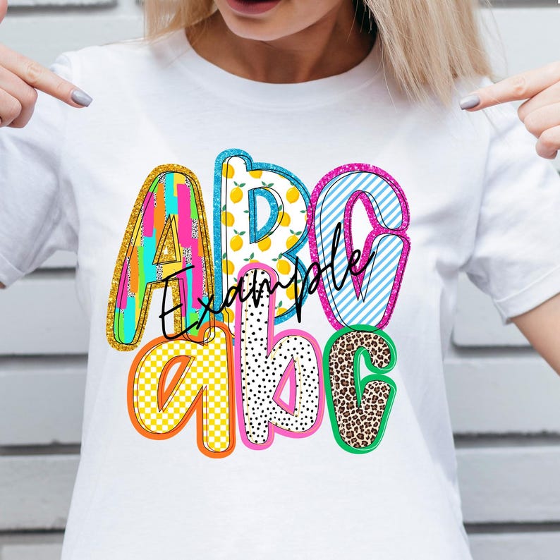 May include: White t-shirt with a colorful ABC design. The letters are filled with various patterns, including stripes, polka dots, and leopard print. The word "Example" is written in cursive across the letters.