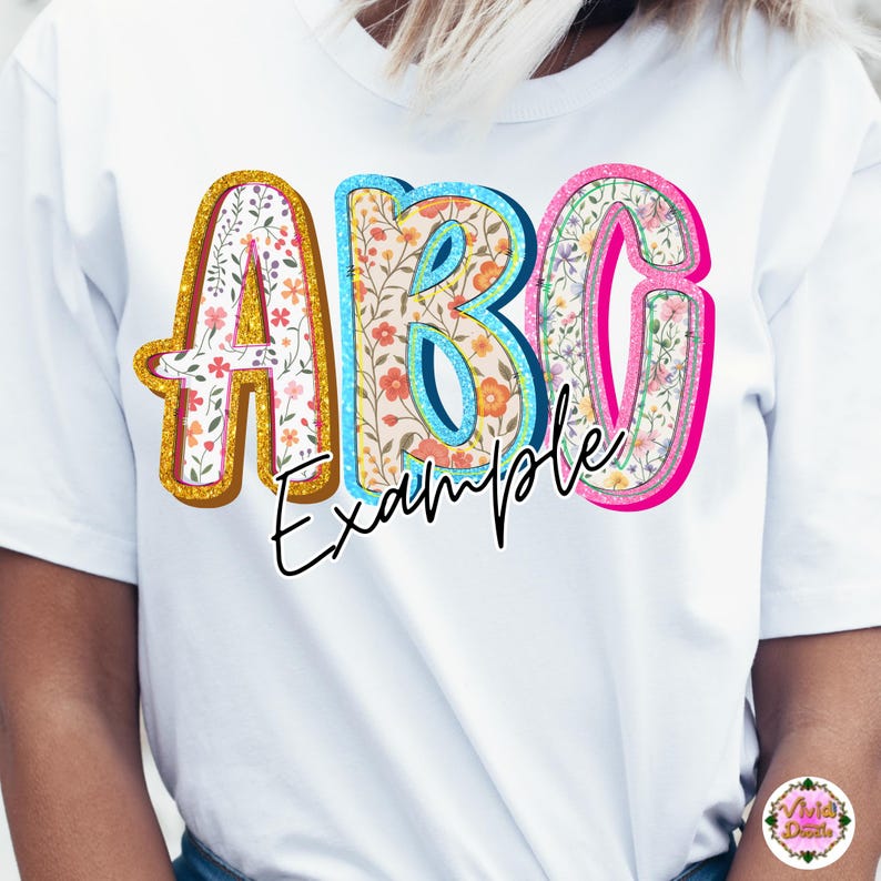 May include: White t-shirt with the word "ABC Example" in a colorful floral design. The letters are outlined in gold, blue, and pink, with a floral pattern inside. The word "Example" is written in black script.
