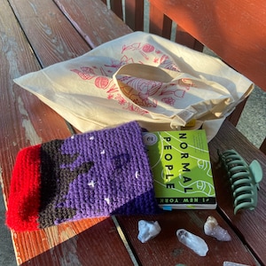 Op de afbeelding: Een paarse en rode gehaakte boekhoes met witte sterren, een boek getiteld "Normal People" van Sally Rooney, een groene haarspeld en verschillende heldere en groene kristallen op een houten bank.