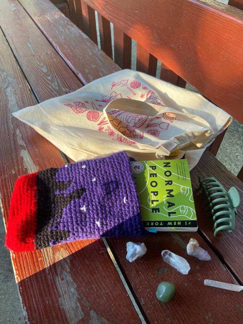 May include: A purple and red crocheted book sleeve with white stars, a book titled "Normal People" by Sally Rooney, a green hair claw, and several clear and green crystals on a wooden bench.