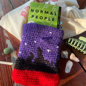 May include: A purple crocheted book sleeve with a black and red design. The sleeve is holding a book titled "Normal People" with the text "#1 NEW YORK TIMES" on the side.
