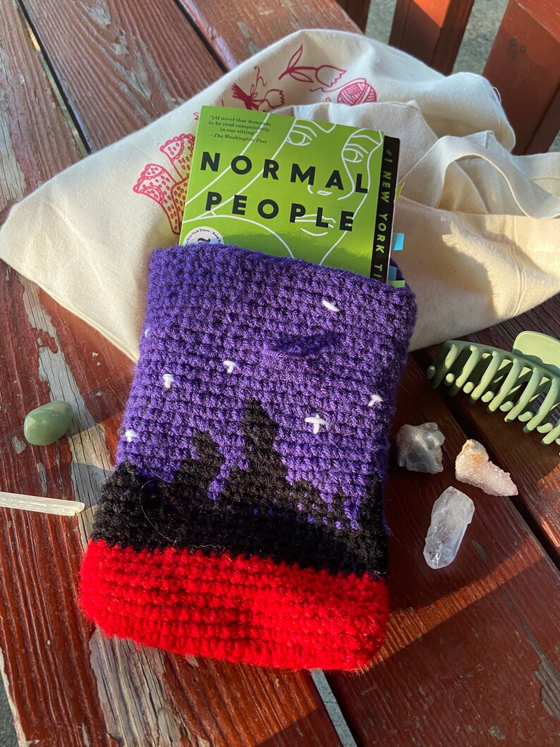 May include: A purple crocheted book sleeve with a black and red design. The sleeve is holding a book titled "Normal People" with the text "#1 NEW YORK TIMES" on the side.
