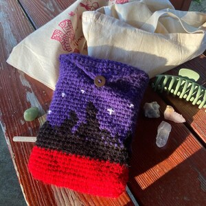 May include: A purple, black, and red crocheted pouch with a button closure. The pouch has a design of a mountain range with stars in the sky.