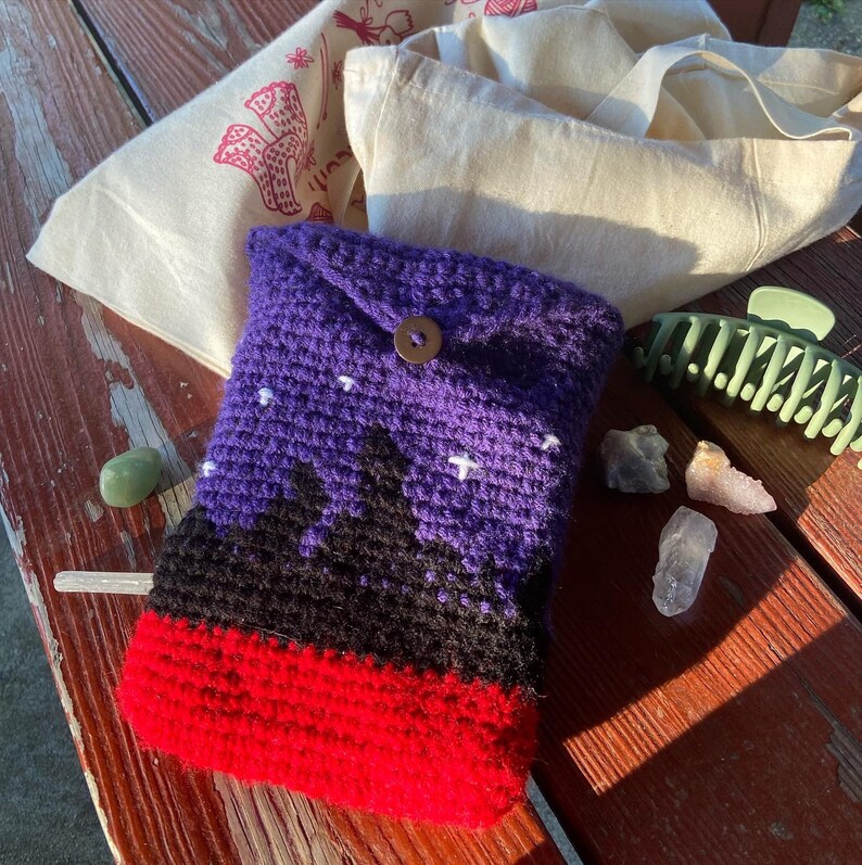 May include: A purple, black, and red crocheted pouch with a button closure. The pouch has a design of a mountain range with stars in the sky.