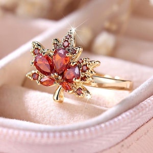 Könnte beinhalten: Ein goldfarbener Ring mit einem Ahornblatt-Design, besetzt mit orange-roten Edelsteinen. Der Ring wird in einer rosa Schmuckschatulle präsentiert, die die feinen Details hervorhebt. Das Design des Rings ist von der Natur inspiriert.