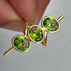 May include: A gold ring featuring three oval-cut green gemstones with a small red gemstone in the center of each. The ring's design is modern and elegant, showcasing the vibrant colors of the gemstones.